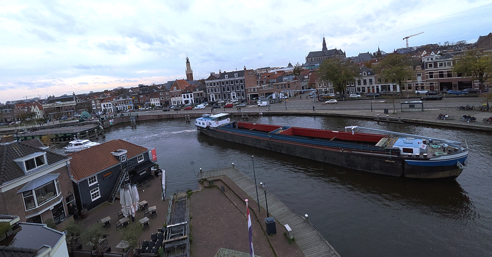 A river bend with a large barge turning the corner in the water. A swing bridge to the left is open. A cityscape is in the background with two church steeples on the horizon.