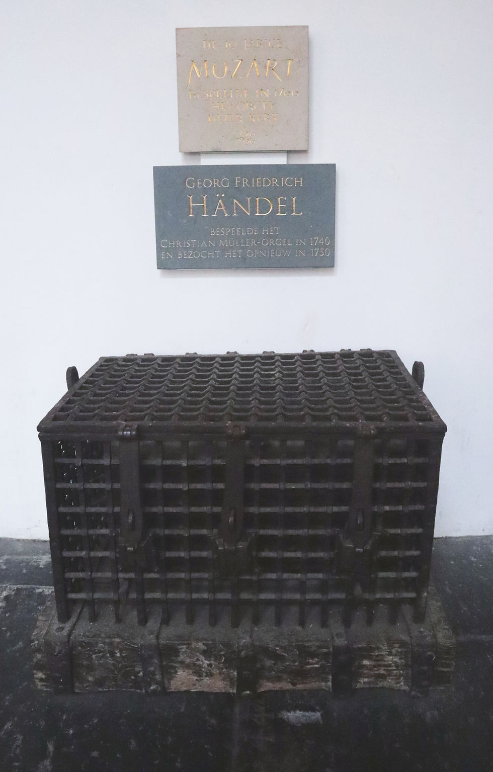 A very old, wrought iron lock box about the size of a treasure chest and two plaques, one reading "Mozart" and the other reading "Handel" on the wall behind the box.