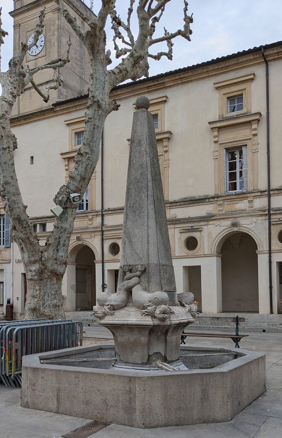 A stone fountain in front of a municipal government building. A small obelisk and four dolphins adorn the fountain.