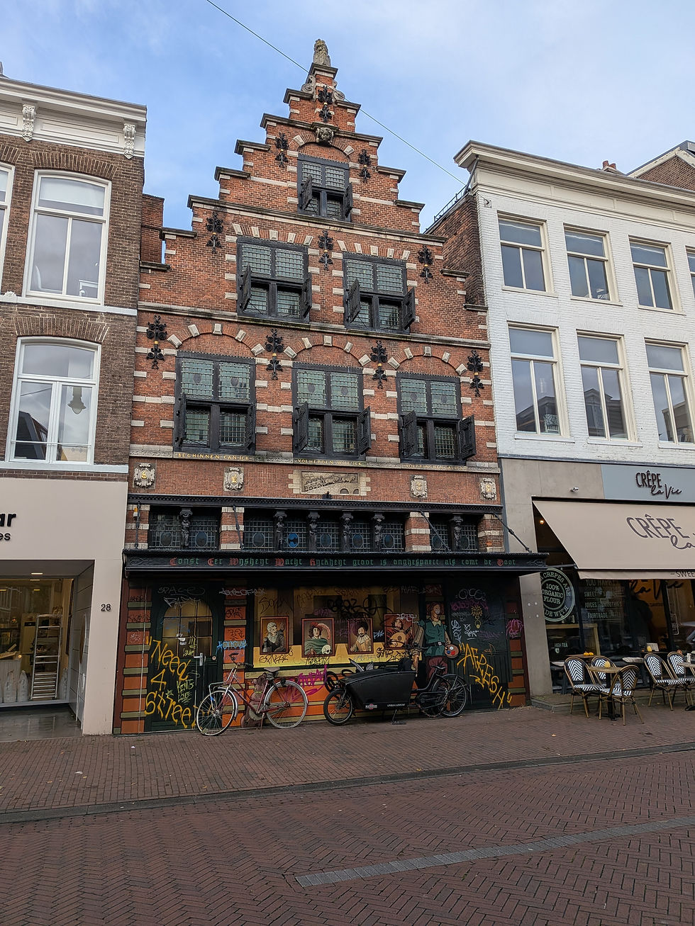 A red brick building with stepped gable facade. It is four stories and has a storefront on the ground level, three windows on the first story, two on the second, and one at the top. It has eight steps to the peaked roof. The road in front is a cobblestone bike lane.