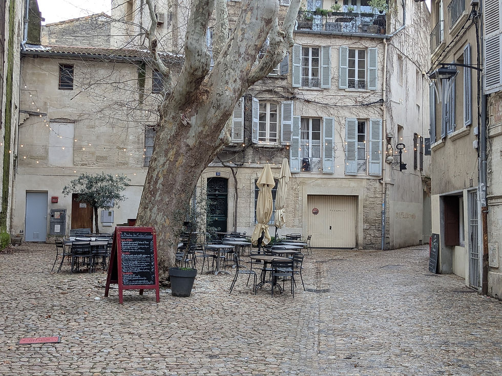 A small cobblestone plaza with a few tables and chalkboard menu for a cafe. A large tree is in the center and a few residential buildings line the plaza.