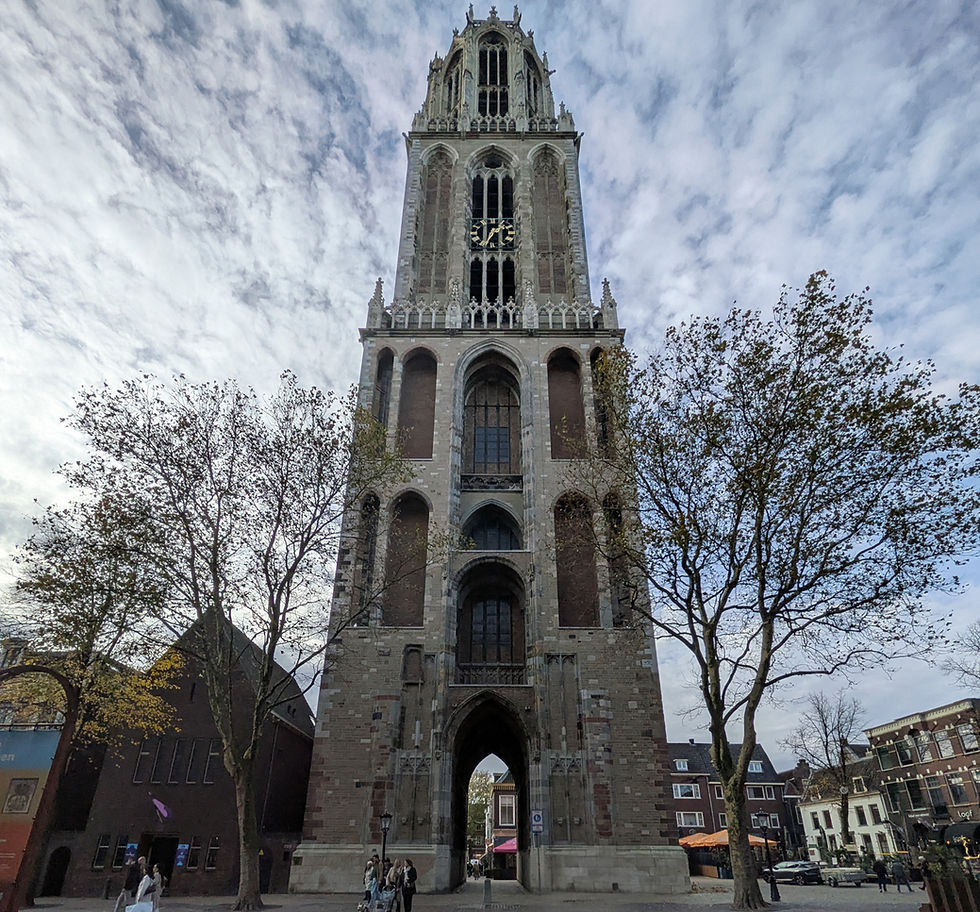 A tall, medieval standalone tower. It has an arched passage through the middle at the bottom, and two arched windows on the two stories about that. There is a clock about 4/5s of the way up. The sky is blue and white with light clouds and the trees have very few leaves.