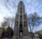A tall, medieval standalone tower. It has an arched passage through the middle at the bottom, and two arched windows on the two stories about that. There is a clock about 4/5s of the way up. The sky is blue and white with light clouds and the trees have very few leaves.