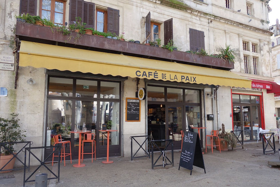 Facade of Cafe de La Paix. The awning is yellow, there are a couple high-top tables outside and a sandwich board menu. Shuttered windows in various states of open and closed are above the awning.