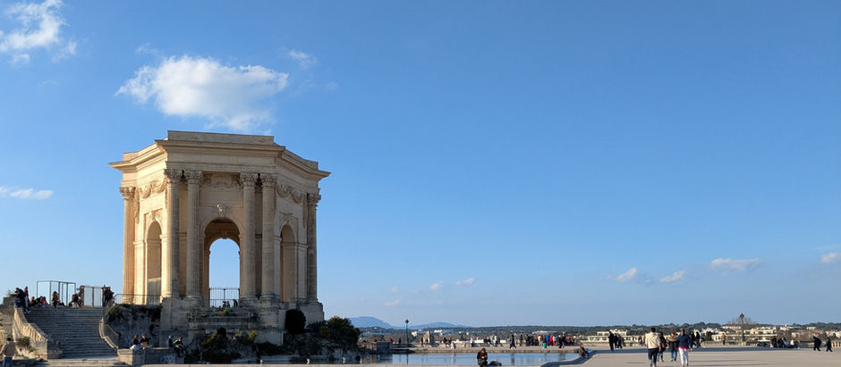 Château d'eau du Peyrou, an arched and columned stone structure overlooking a pond on a plaza. People sit nearby and walk by. Mountains are on the distant horizon.