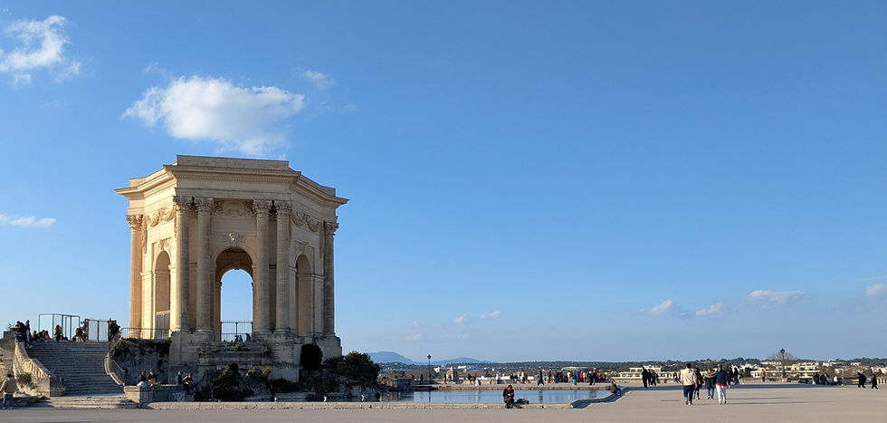 Château d'eau du Peyrou, an arched and columned stone structure overlooking a pond on a plaza. People sit nearby and walk by. Mountains are on the distant horizon.
