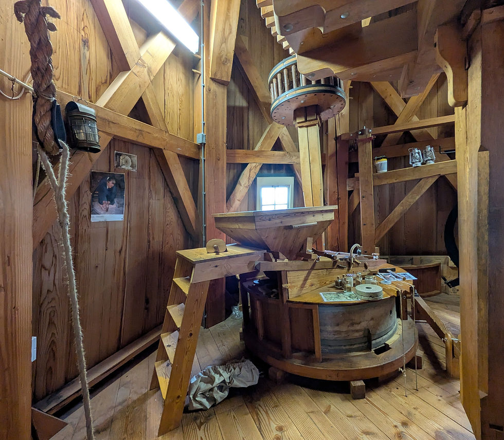 A small wooden room with gears, bins, ropes and wooden stairs. It is the inside of a windmill.