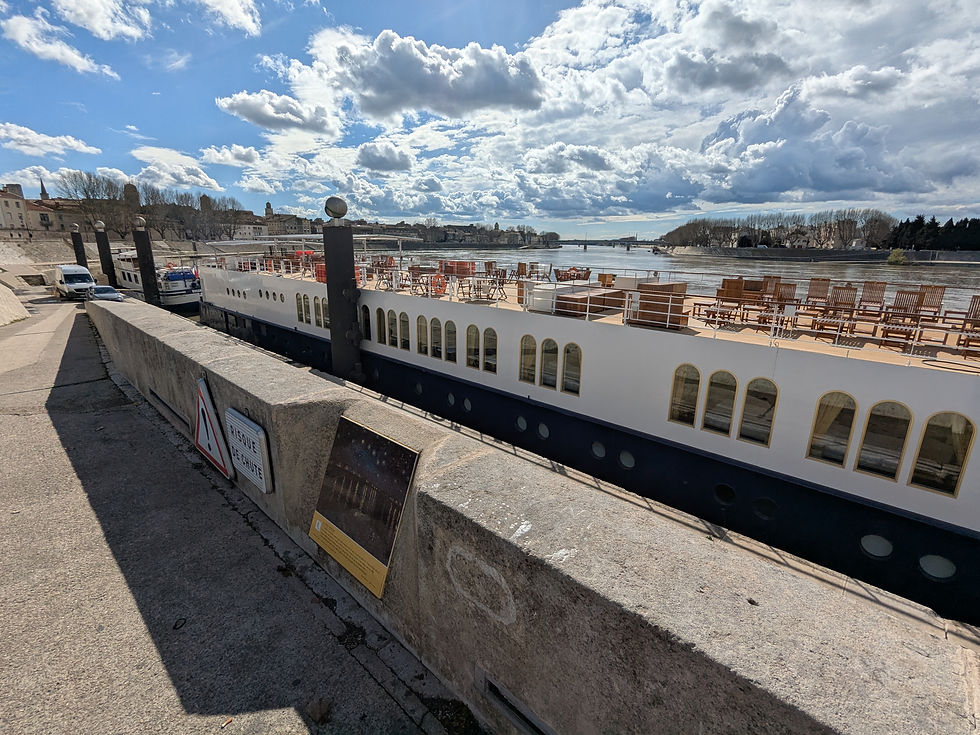 A guay along a river. The cement wall has an easel depicting van Gogh's La Nuit Etoilee. There's a long tour boat in the water. The sky is blue with many puffy, white clouds.