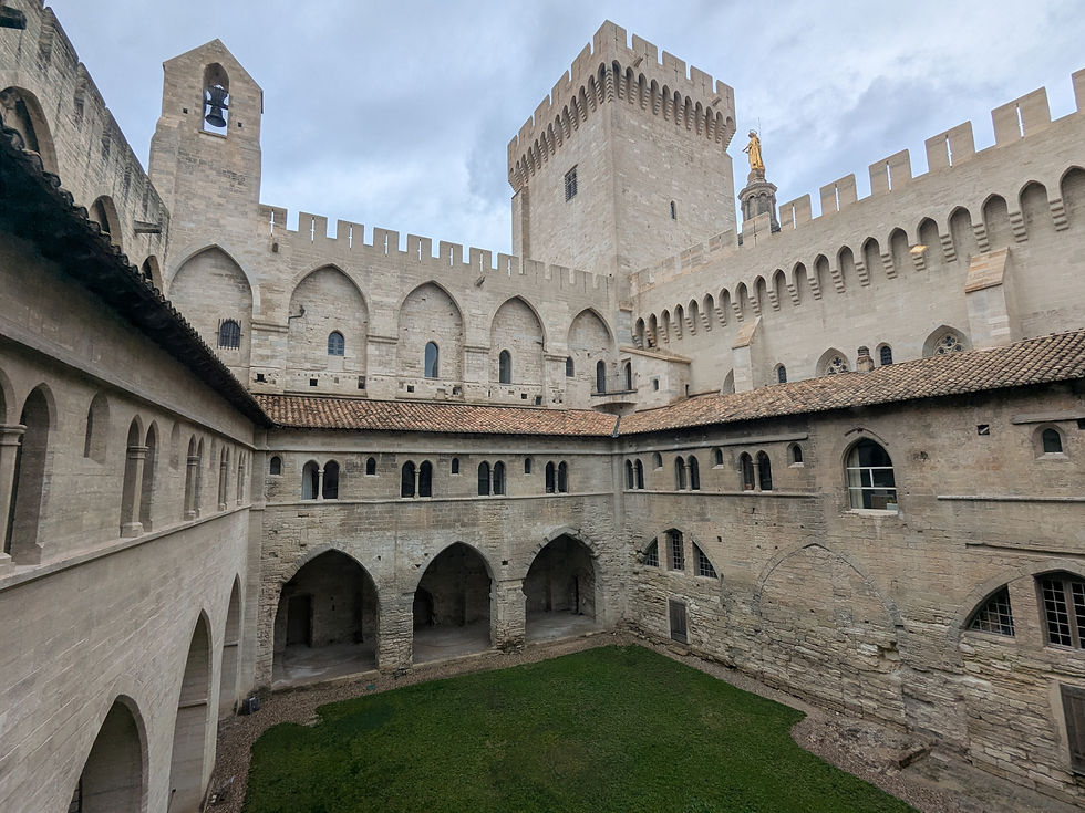 Courtyard of a palace, with towers, ramparts, church steeple (with golden statue of Mary), a bell tower, archways and green grass visible.