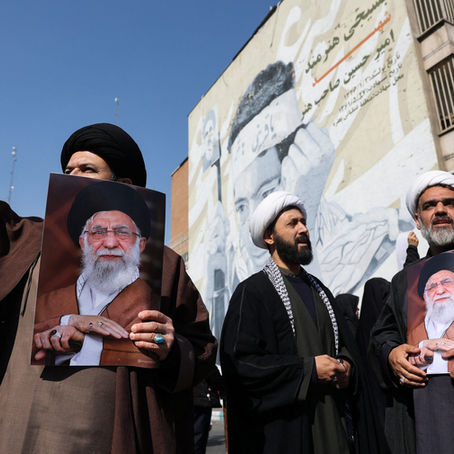 Three Shia clerics stand outdoors during a demonstration, two holding posters of Iran’s supreme leader while one raises his fist; a large mural of the same leader is painted on a building behind them.