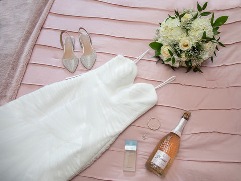 Flat lay of bridal items. Wedding dress, shoes, bouquet, perfume, jewelry and a bottle of champagne on a pink background.