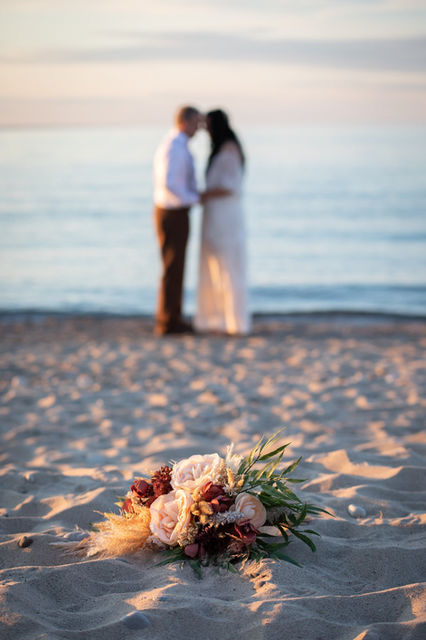 a bride and groom holding hands standing by the water on the beach and blurred in the background, the bridal bouquet is in the foreground on the sand and in focus