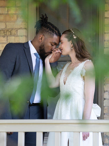 A man and a woman standing on a porch by a white railing, he is holding her hand and kissing it.