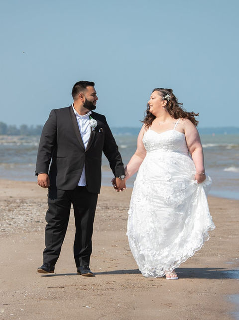 A bride and groom walking on a sandy beach by the edge of the lake holding hands.