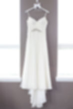 The white wedding dress is hanging from a personalized "bride" hanger on the wall between two windows