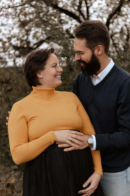 Babybauch und Familienfotos Olching. Eine schwangere Frau und ihr Mann stehen vor einem Feld. Beide haben ihre Hand oben auf den Bauch gelegt und schauen sich lachend an