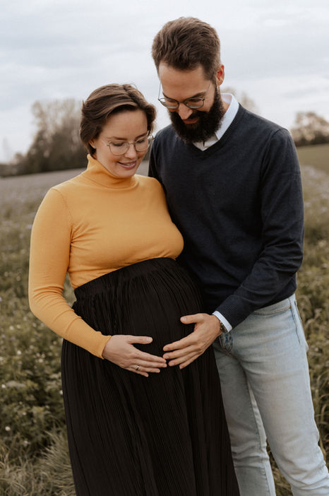 Babybauch und Familienfotos Olching. Eine schwangere Frau und ihr Mann stehen vor einem Feld. Beide haben ihre Hand auf den Bauch gelegt und schauen den Bauch an