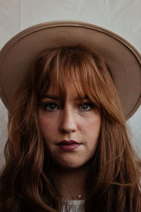 Foto van het gezicht van een vrouw met oranje haar en een grote hoed op.