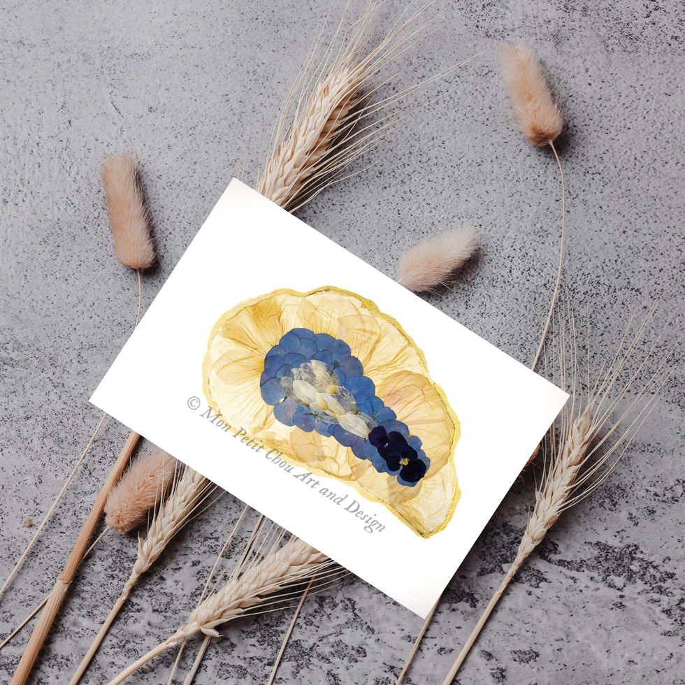 Pressed flower oyster art greeting card with blue petals on a golden shell shape, styled with dried grasses on a stone surfac