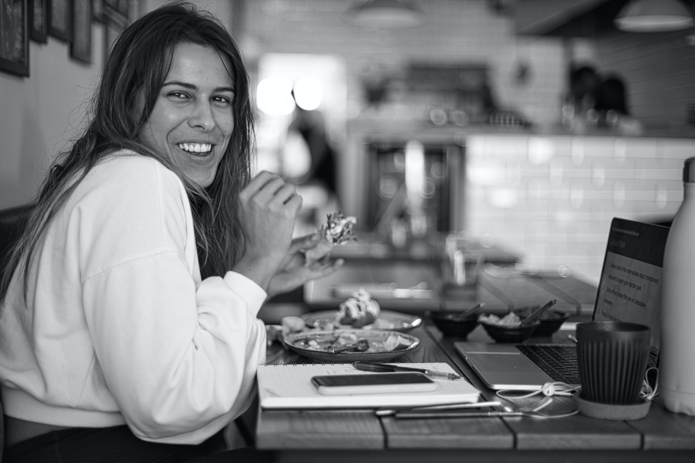 smiling woman at restaurant table with food, laptop, phone and notebook