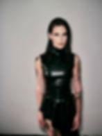 DJ Rebekah with wet hair in a black leather outfit stands against a plain background, showing tattoos with a confident expression.