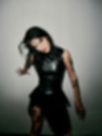 DJ and producer Rebekah with wet hair in black leather dress poses against a gray wall, showing tattoos. Moody, intense expression.