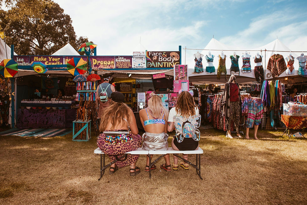 Three people sit on a bench at a colorful festival market with clothes and umbrella displays under a clear blue sky. Signs read "Face & Body Painting."