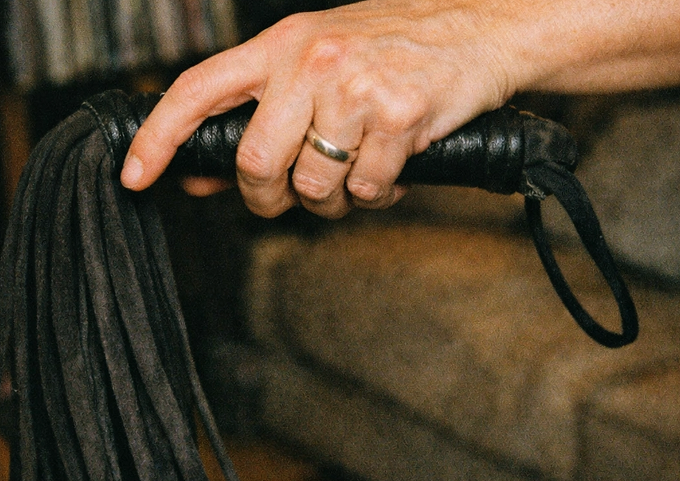 A hand with a silver ring holds a black leather flogger against a blurred background, suggesting a domestic setting. The mood is subtle.