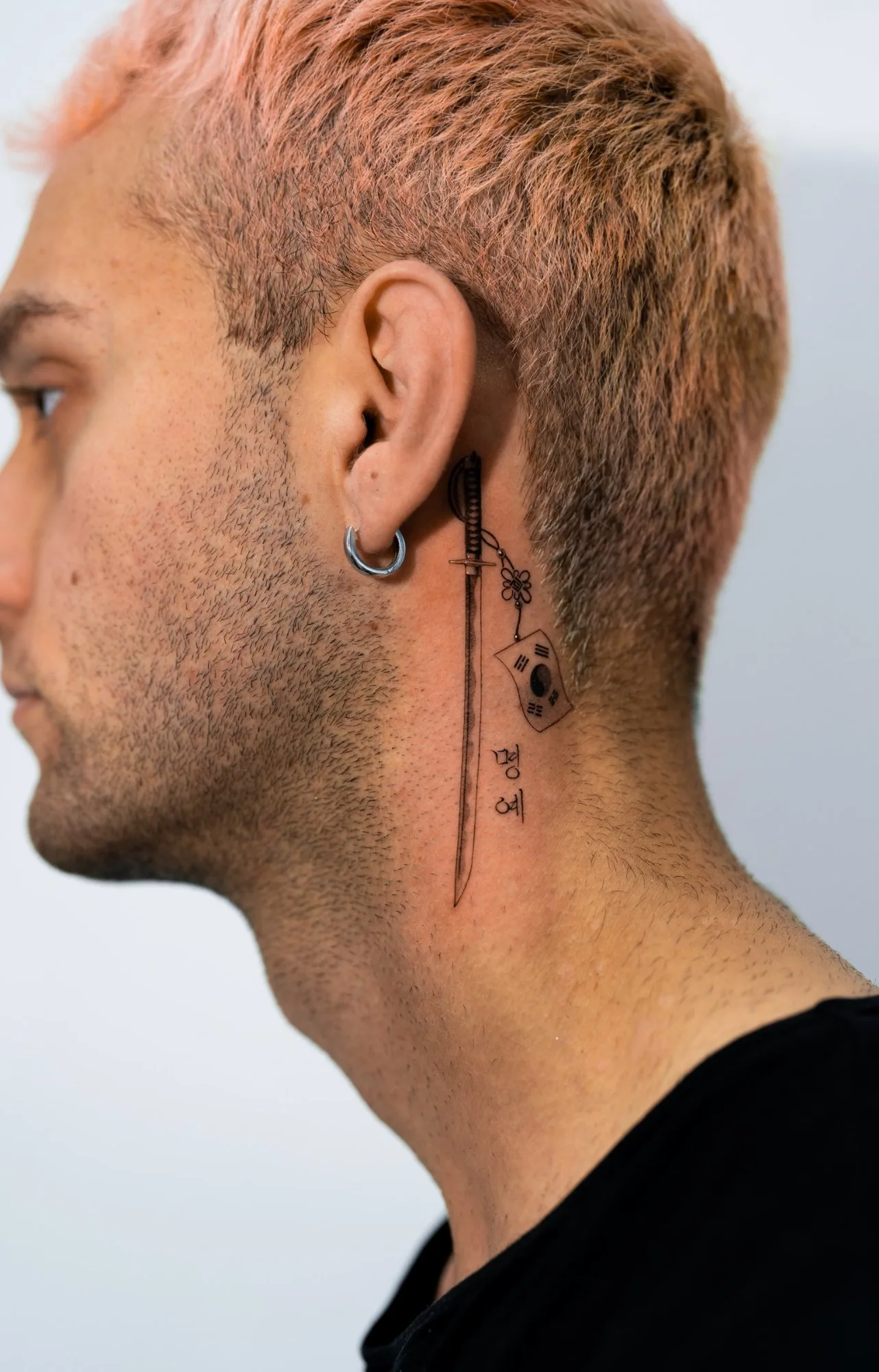 Fineline tattoo behind the ear featuring a vertical katana sword with a small Korean Taegukgi flag charm and Korean Hangul script below it on the neck. Seoul Ink Tattoo, Gangnam, Seoul, South Korea.