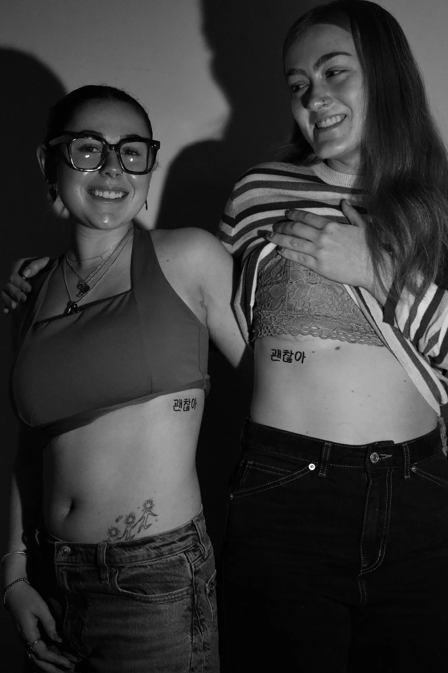 Two smiling friends showing matching Korean Hangul script tattoos reading '괜찮아' (meaning 'it's okay') on each of their lower ribs/sides. Seoul Ink Tattoo, Gangnam, Seoul, South Korea.