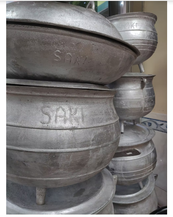 Stacked silver metal pots and lids in a kitchen corner, some embossed with SANI, showing a worn, utilitarian look.