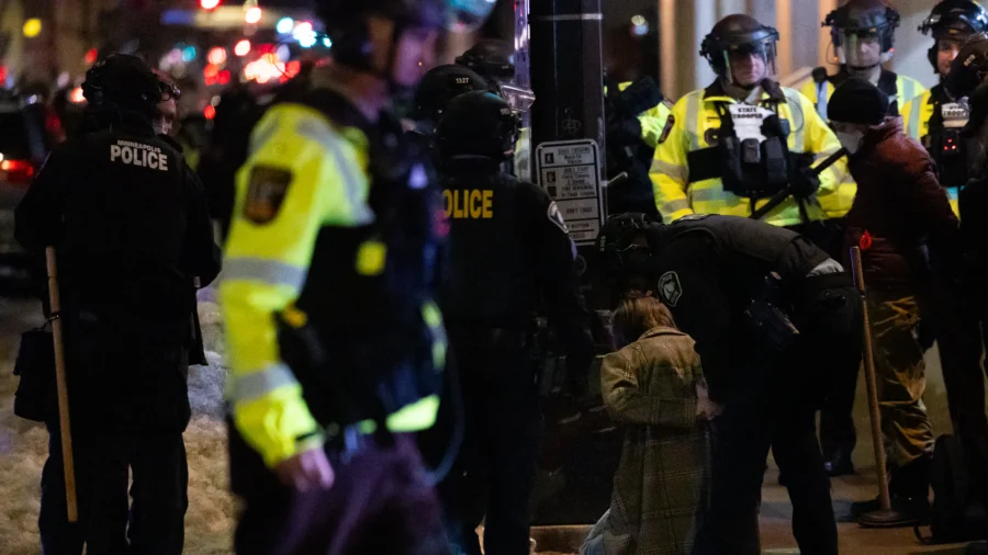 Minneapolis police officers in riot gear and high-visibility yellow vests surround a kneeling or detained protester on a snowy downtown street at night during the January 10, 2026, anti-ICE protests. Officers hold batons while one assists or restrains the person on the ground amid the unlawful assembly response.