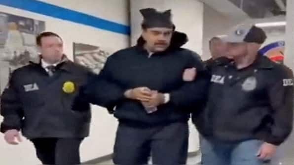 Nicolás Maduro, former Venezuelan president, is escorted in handcuffs by two DEA agents through a hallway (likely at DEA headquarters in New York). He wears a black hooded sweatshirt, appears blindfolded or hooded, and holds a plastic water bottle in his cuffed hands. The agents are in dark DEA jackets with visible badges. Scene from his January 2026 U.S. custody transfer.