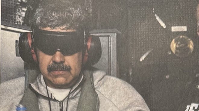 Nicolás Maduro, the former president of Venezuela, is shown in U.S. custody aboard the USS Iwo Jima following his capture. He is wearing a gray sweatshirt and sweatpants, large black sound-blocking headphones (ear protectors), a dark blindfold covering his eyes, and is handcuffed. He holds a plastic bottle of water in his hands. The dimly lit photo captures him seated in what appears to be a military or naval environment, with part of another person's shoulder or gear visible on the right side.