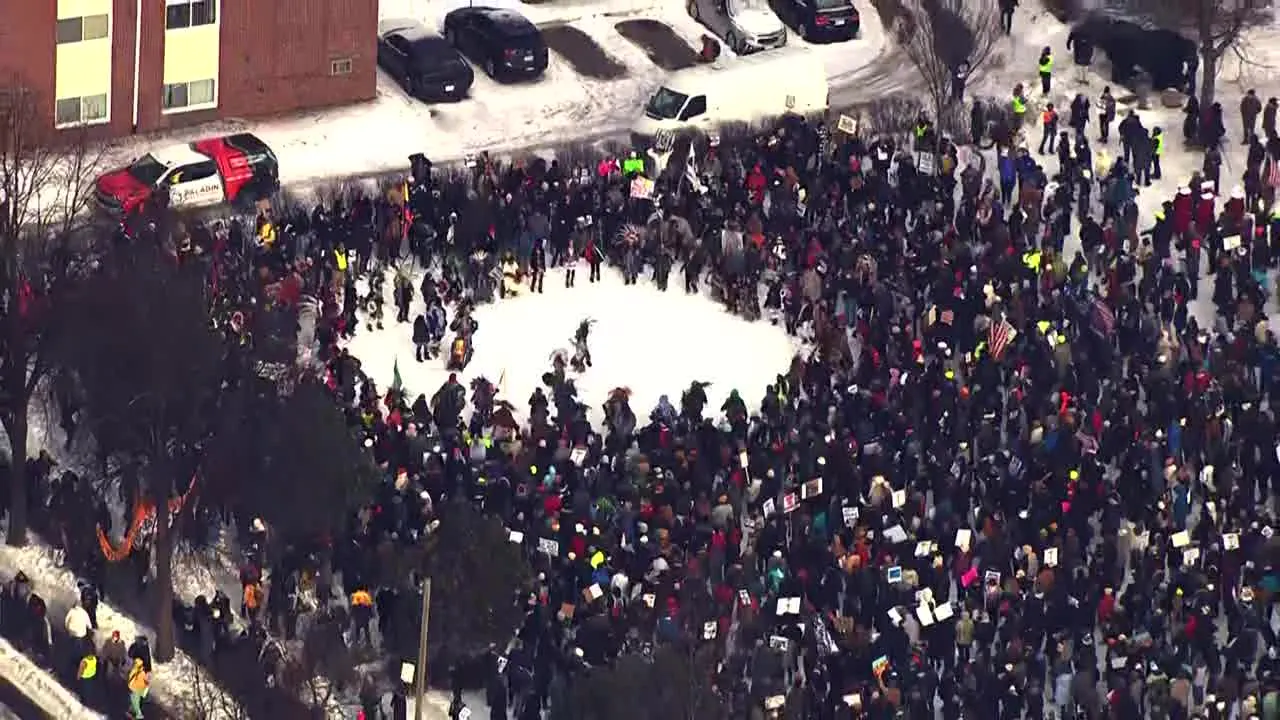 Aerial drone view of a massive crowd of anti-ICE protesters in Minneapolis on a snowy January day in 2026, forming a large heart shape in the snow-covered street or park. Hundreds of people in winter coats hold colorful signs, flags, and banners while standing in a circular formation around the center, with parked cars, trees, and buildings visible in the background.