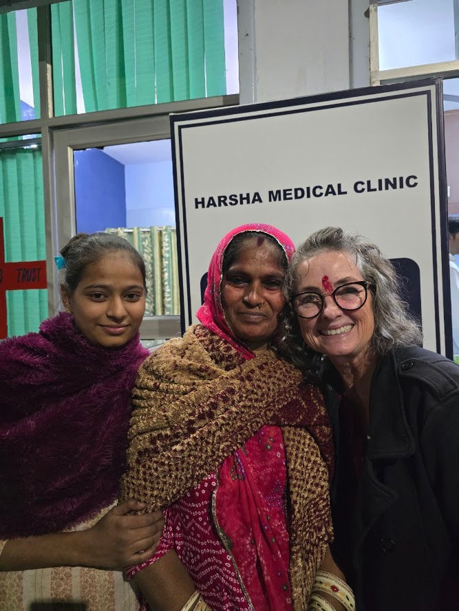 Three smiling women stand close together in front of a "Harsha Medical Clinic" sign. The mood is warm and joyful.
