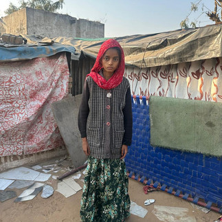 Young girl in red scarf looks sad and ill. She is standing in a dirt courtyard with two tents behind