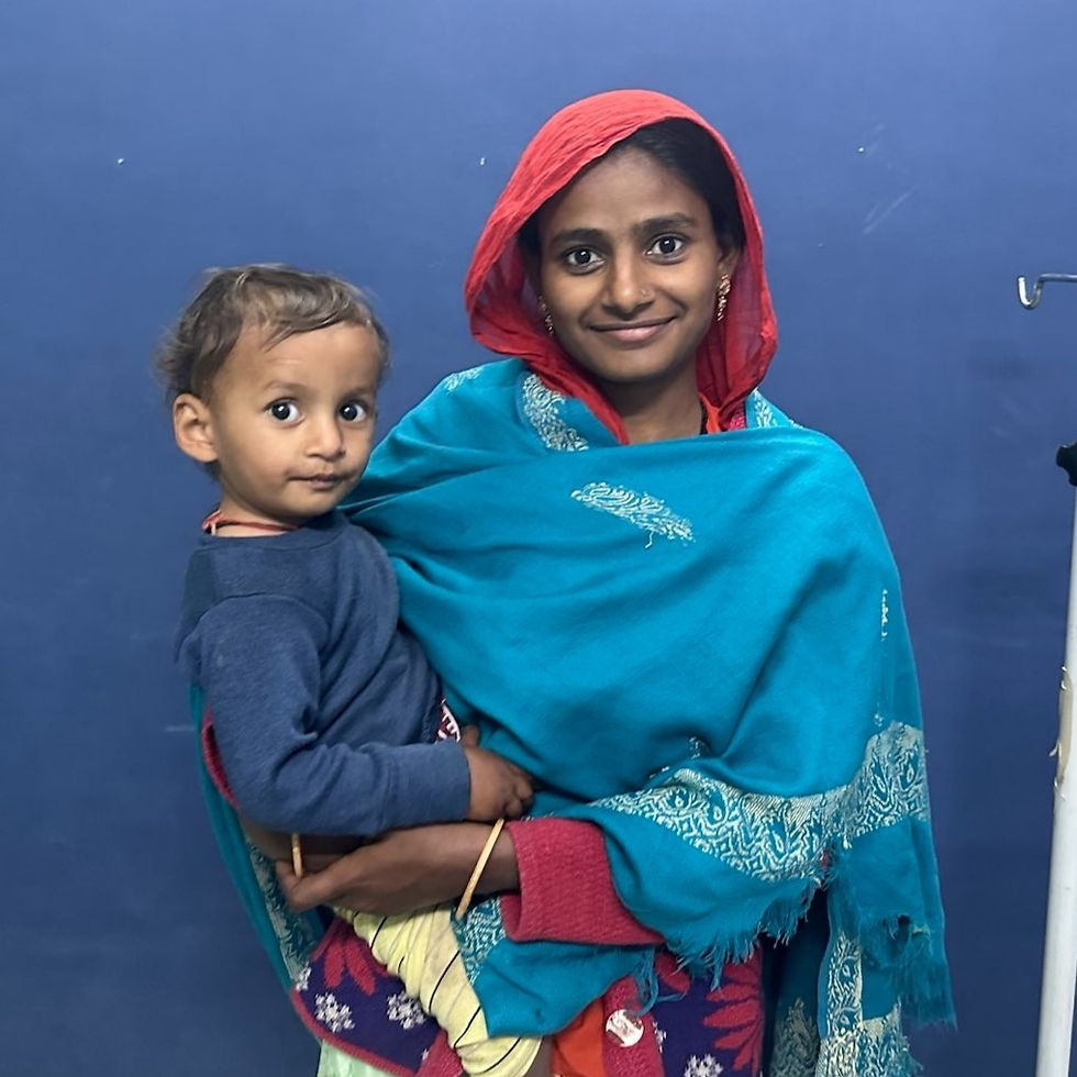 A woman in a blue shawl and red headscarf holds a young child against a blue wall in a clinic. Both are smiling, creating a warm, joyful mood.
