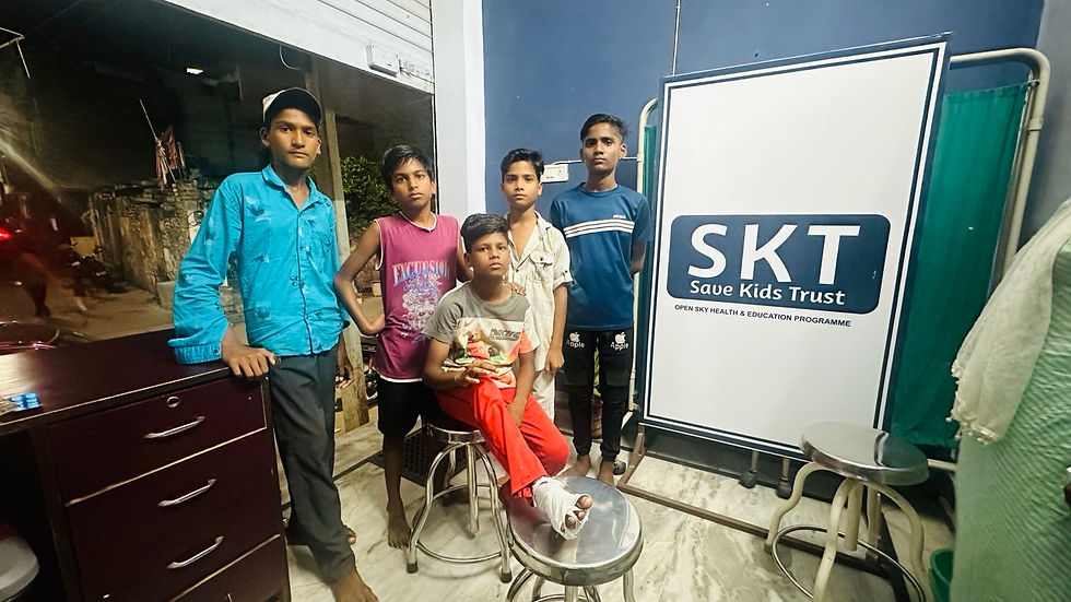 Five boys in casual attire stand indoors. One sits on a stool with a bandaged foot. "SKT Save Kids Trust" board visible. Evening setting.