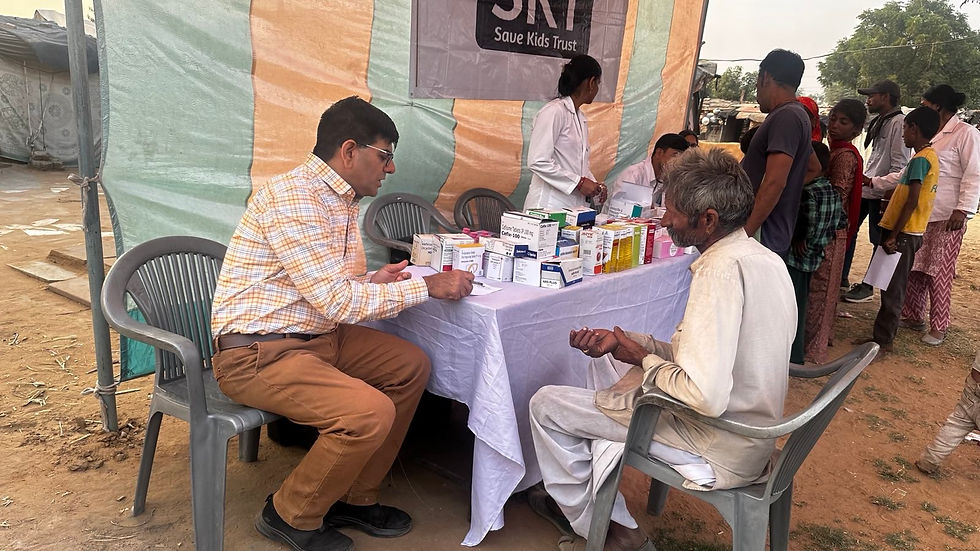 Dr. Sharwan Saini talking to elderly man at a medical camp table outdoors. Medicines displayed; people waiting in line. Banner reads "Save Kids Trust."