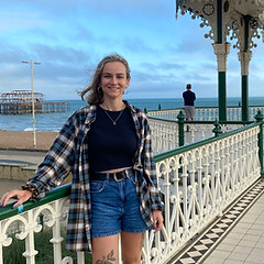 Angelika is standing by a railing, with one of her hands on the railing. Her hair is shoulder length and wind-swept. She is wearing a black crop top, with blue jean shorts and a checkered shirt. Tattoos are visible on her legs. 