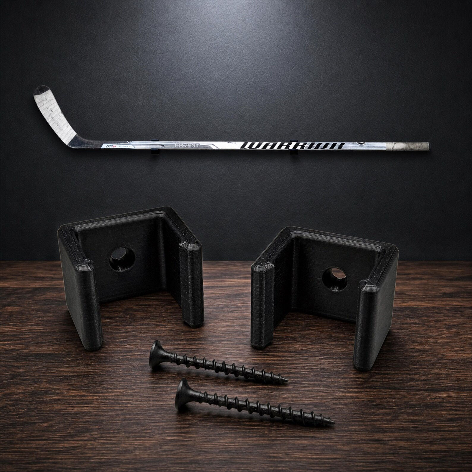 Two black U-shaped plastic wall mounts for a hockey stick, accompanied by two black screws. The mounts feature a central circular hole for installation, designed to hold a Warrior brand hockey stick horizontally against a wall.