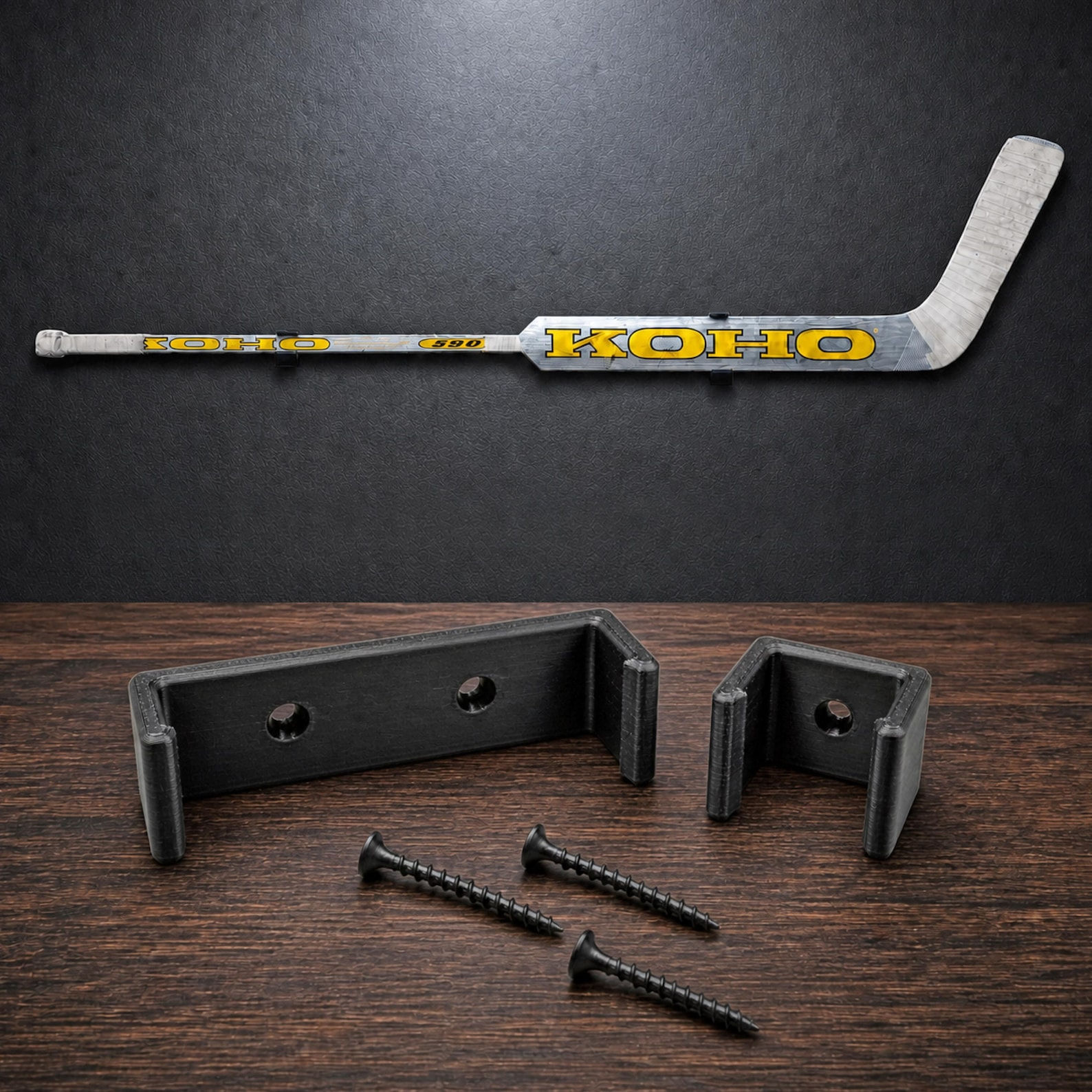 A silver and yellow KOHO 590 ice hockey stick mounted horizontally above two black U-shaped plastic wall brackets and three black metal screws. The stick features the brand name KOHO in yellow text with a black outline.