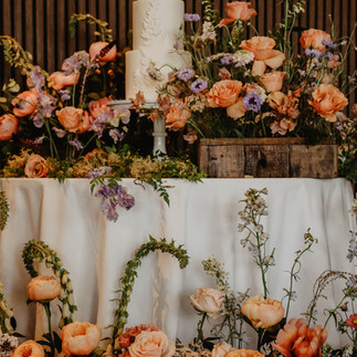 a white three tiered cake with white wildflower detailing, stood upon a white cake stand. The cake table is adorned with an abundance of wildflower arrangements.