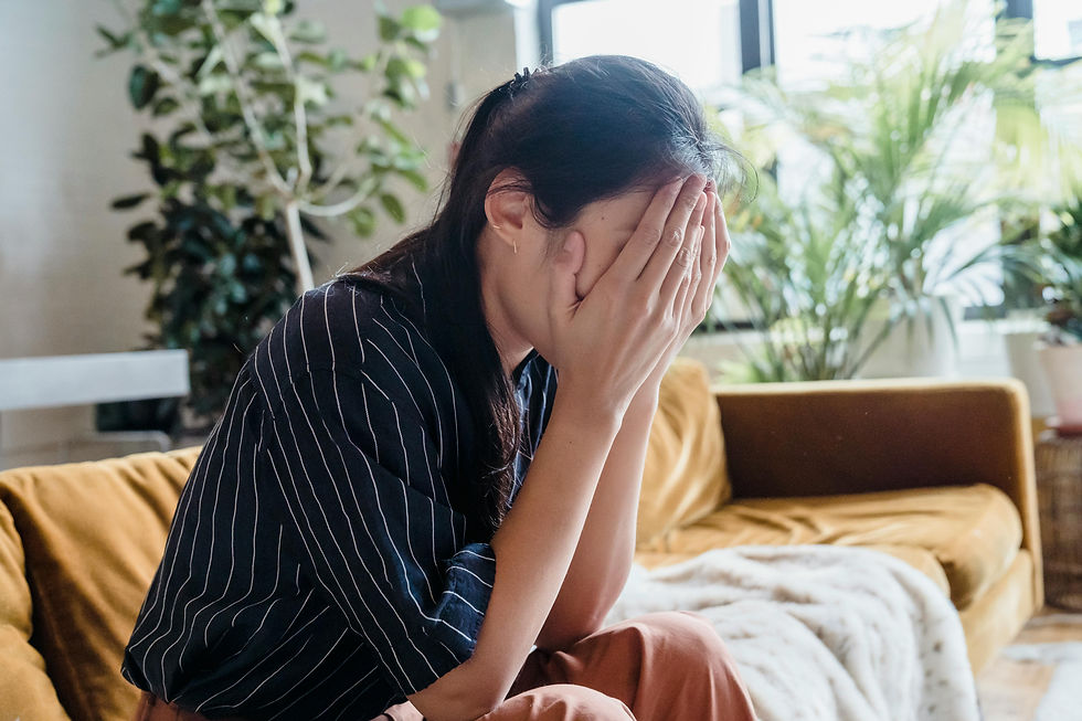 Woman in striped shirt sits on sofa, covering face with hands, appearing upset. Indoor setting with plants and a window in the background.