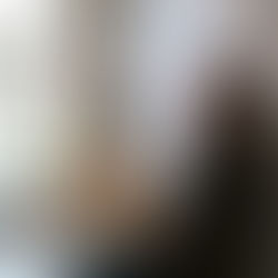 A woman in a hoodie sits in a kitchen, gazing thoughtfully. Bright window, plants, and table with flowers enhance the serene mood.