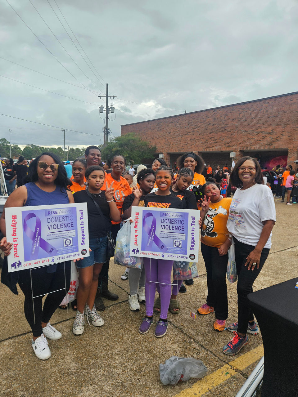 Candy Christophe, Interim Executive Director of Re-Entry Solutions, stands with a group of women and children during the Southern Girl Ryderz & Southern Ryderz 1st Annual Trunk or Treat in Alexandria, LA. The group smiles while holding RISE Above Domestic Violence awareness signs promoting support and resources for families.