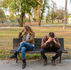 A man and woman sit on a park bench, visibly upset. The woman faces away, and the man covers his face. Trees and a building are in the background.