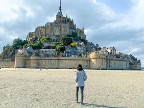 Person in a gray jacket walking on sandy beach toward Mont-Saint-Michel, a historic island with stone buildings, under a cloudy sky. Mont Saint-Michel, Normandy, France. Pilgrimage