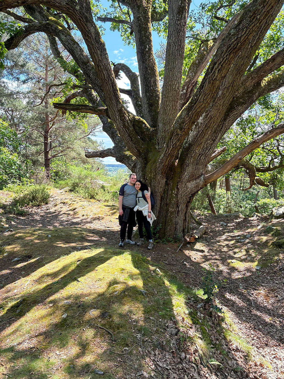 Smiling couple stands under a huge old tree on a sunny forest trail, with mossy ground and blue sky overhead. Bouquet de la Reine Amélie Fontainebleau Forest Paris France